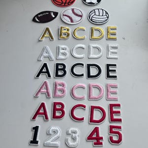 May include: A set of iron-on patches featuring letters A through E in gold, white, black, pink, and red, as well as numbers 1 through 9 in white, black, gold, and pink. There are also patches of a basketball, football, baseball, and volleyball.