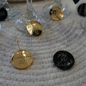 May include: A collection of wine glass charms. Some are gold-colored, some are silver, and some are black. The charms have names and dates engraved on them, including "Friendsgiving 2025" and "Miss XV Delilah".