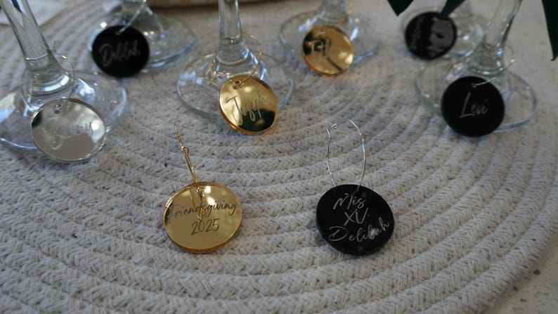 May include: A collection of wine glass charms. Some are gold-colored, some are silver, and some are black. The charms have names and dates engraved on them, including "Friendsgiving 2025" and "Miss XV Delilah".