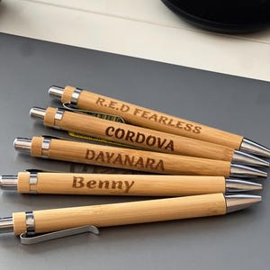 May include: A collection of five bamboo pens with silver accents. Each pen is engraved with a different name or phrase: "R.E.D FEARLESS", "CORDOVA", "DAYANARA", and "Benny". The pens are arranged on a gray surface.