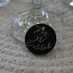 May include: A black, round wine glass charm with the words "Mis XV Delilah" in white script. The charm is attached to a wine glass. The background is a white, textured surface.