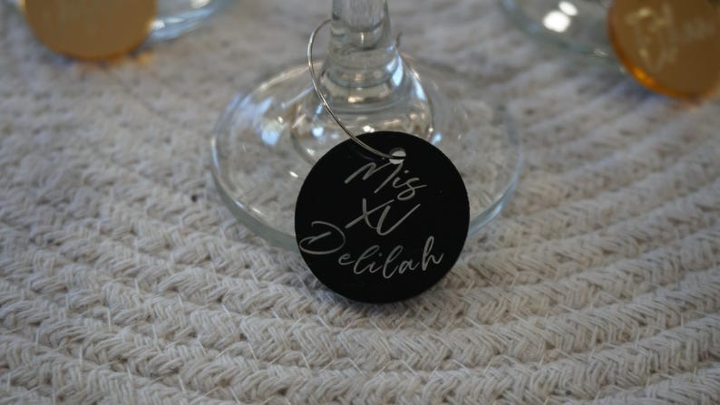 May include: A black, round wine glass charm with the words "Mis XV Delilah" in white script. The charm is attached to a wine glass. The background is a white, textured surface.