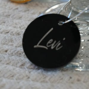 May include: A black, round tag with the name "Levi" in white script. The tag is attached to a clear glass object, possibly a wine glass, and rests on a textured white surface. The tag is likely a place setting or gift tag.