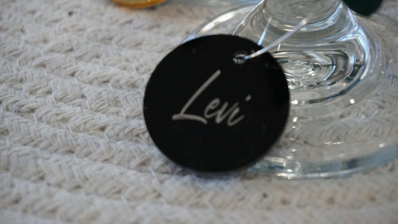 May include: A black, round tag with the name "Levi" in white script. The tag is attached to a clear glass object, possibly a wine glass, and rests on a textured white surface. The tag is likely a place setting or gift tag.