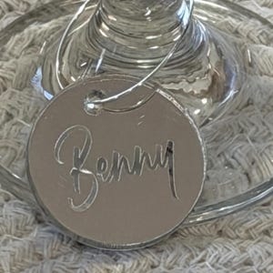 May include: A silver-colored wine glass charm with the name "Benny" cut out. The charm is round and attached to a wine glass. The background is a light-colored, textured surface.