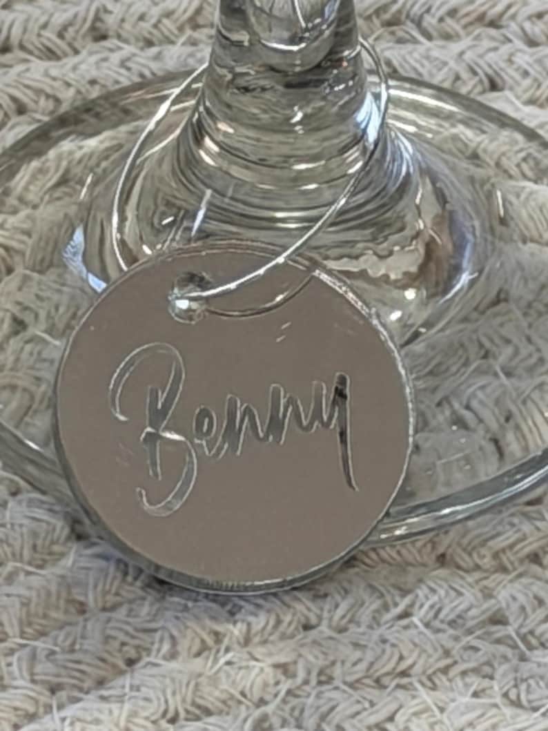 May include: A silver-colored wine glass charm with the name "Benny" cut out. The charm is round and attached to a wine glass. The background is a light-colored, textured surface.