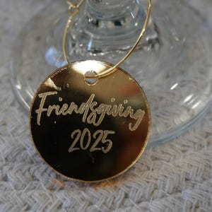 May include: A gold-colored Friendsgiving 2025 wine charm. The charm is round and hangs from a thin gold wire. The text "Friendsgiving 2025" is etched in a cursive font. The charm is resting on a clear wine glass.