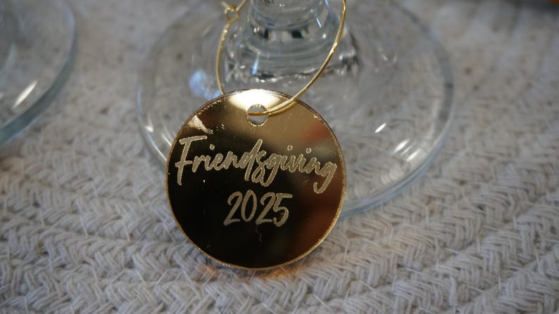 May include: A gold-colored Friendsgiving 2025 wine charm. The charm is round and hangs from a thin gold wire. The text "Friendsgiving 2025" is etched in a cursive font. The charm is resting on a clear wine glass.
