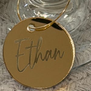 May include: A gold-colored, circular wine charm with the name "Ethan" engraved in a cursive font. The charm is attached to a thin, gold-colored wire loop, designed to hang on a wine glass. The background is blurred, suggesting a party or event setting.