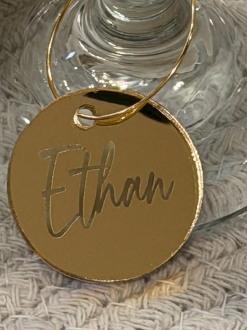 May include: A gold-colored, circular wine charm with the name "Ethan" engraved in a cursive font. The charm is attached to a thin, gold-colored wire loop, designed to hang on a wine glass. The background is blurred, suggesting a party or event setting.