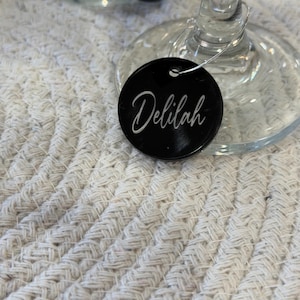 May include: A close-up of a wine glass with a black circular tag that reads "Delilah" in white script. The tag is attached to the glass with a silver wire. The background is a white, textured surface.