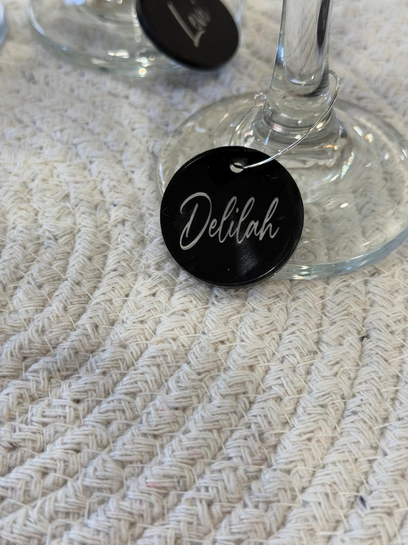 May include: A close-up of a wine glass with a black circular tag that reads "Delilah" in white script. The tag is attached to the glass with a silver wire. The background is a white, textured surface.