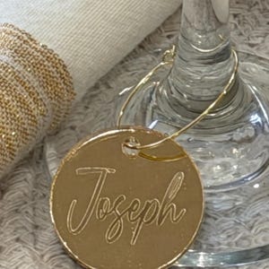 May include: A gold-colored, circular wine charm with the name "Joseph" engraved in a cursive font. The charm is attached to a thin, gold-colored wire that loops around a wine glass. The background includes a white napkin and a clear wine glass.
