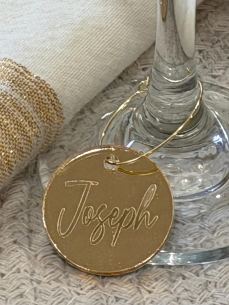 May include: A gold-colored, circular wine charm with the name "Joseph" engraved in a cursive font. The charm is attached to a thin, gold-colored wire that loops around a wine glass. The background includes a white napkin and a clear wine glass.