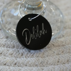 May include: A black wine glass charm with the name "Delilah" in elegant cursive script. The charm is round and attached to a silver wire, resting on a wine glass. The background is a textured, off-white surface.