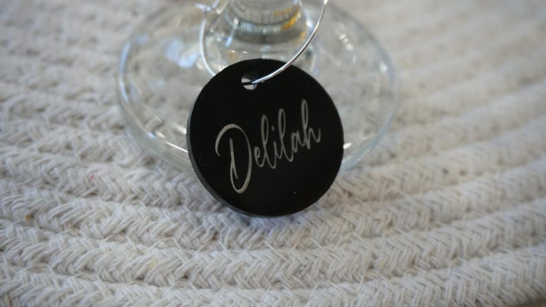 May include: A black wine glass charm with the name "Delilah" in elegant cursive script. The charm is round and attached to a silver wire, resting on a wine glass. The background is a textured, off-white surface.
