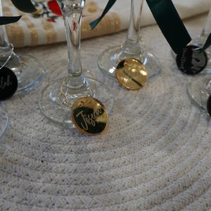 May include: Close-up of several wine glasses with personalized name tags. The tags are round or oval, in silver, gold, and black, with names like "Debbie" and "Tiffany" engraved. Each glass has a dark green ribbon tied around the stem.
