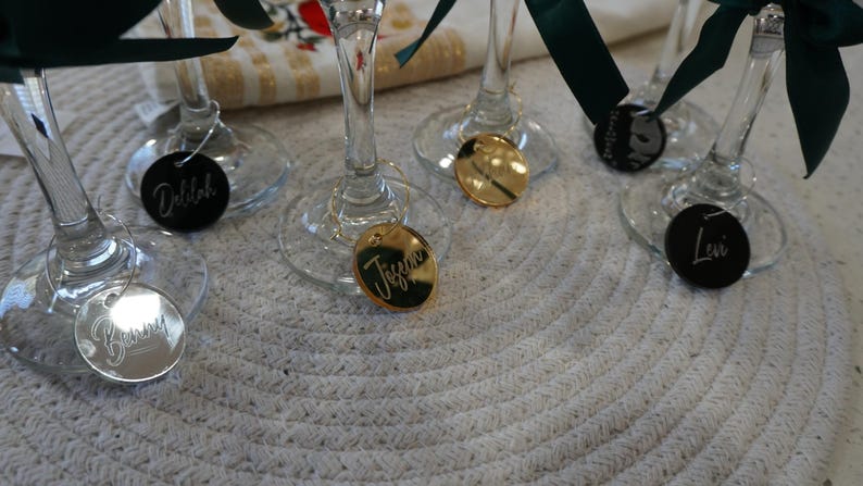 May include: Close-up of several wine glasses with personalized name tags. The tags are round or oval, in silver, gold, and black, with names like "Debbie" and "Tiffany" engraved. Each glass has a dark green ribbon tied around the stem.