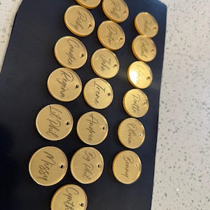 May include: A collection of gold-colored, circular acrylic name tags with engraved names. Each tag has a small hole for attachment. Names include Fiona, Ricky, Candice, Regina, Lil Phil, Missy, Cynthia, Rob, Deuter, John, Irene, Andree, Big Phil, Jackie, Callah, Vince, Pottie, Olivia, and Benny.