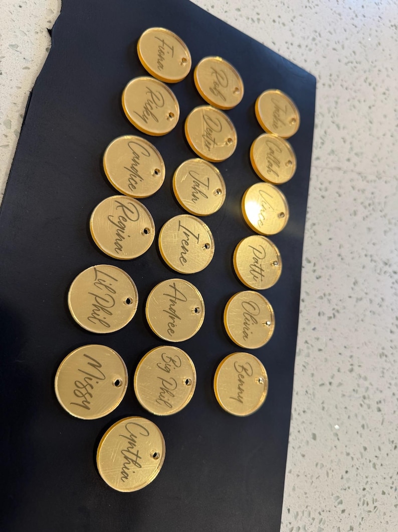 May include: A collection of gold-colored, circular acrylic name tags with engraved names. Each tag has a small hole for attachment. Names include Fiona, Ricky, Candice, Regina, Lil Phil, Missy, Cynthia, Rob, Deuter, John, Irene, Andree, Big Phil, Jackie, Callah, Vince, Pottie, Olivia, and Benny.