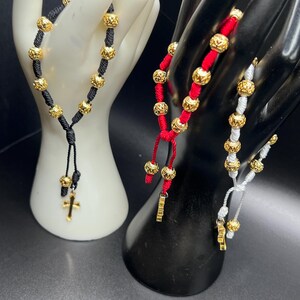 May include: Three bracelets with gold beads and different colored cords. The bracelets are black, red, and white. Each bracelet has a gold charm at the end.