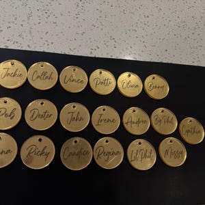 May include: A collection of gold-colored, circular name tags with individual names engraved in a cursive font. The tags are arranged on a black surface. Each tag has a small hole at the top for attachment.
