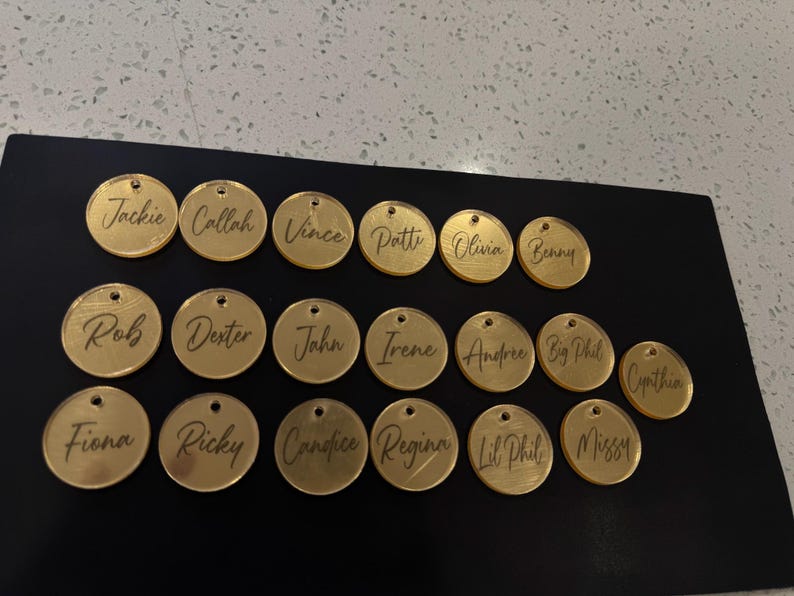 May include: A collection of gold-colored, circular name tags with individual names engraved in a cursive font. The tags are arranged on a black surface. Each tag has a small hole at the top for attachment.