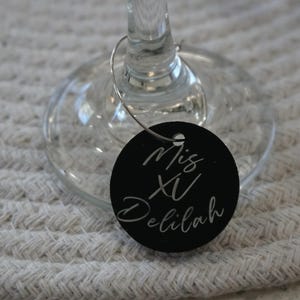 May include: A black wine glass charm with the text "Mis XV Delilah" in white script. The charm is round and attached to a wine glass. The background is a white woven placemat. The charm is a decorative item.