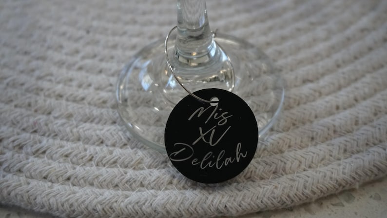 May include: A black wine glass charm with the text "Mis XV Delilah" in white script. The charm is round and attached to a wine glass. The background is a white woven placemat. The charm is a decorative item.