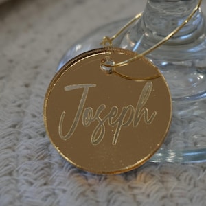 May include: A gold-colored, circular place card with the name "Joseph" engraved in a cursive font. The place card is attached to a wine glass with a thin, gold-colored wire. The background is a white, textured surface.