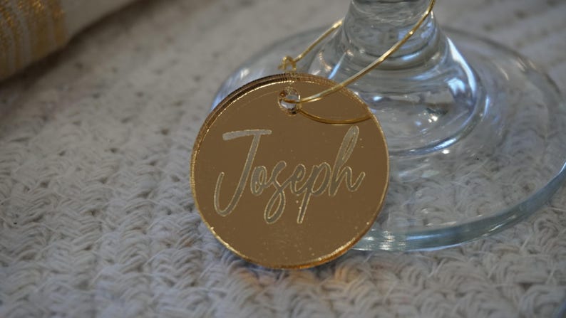 May include: A gold-colored, circular place card with the name "Joseph" engraved in a cursive font. The place card is attached to a wine glass with a thin, gold-colored wire. The background is a white, textured surface.