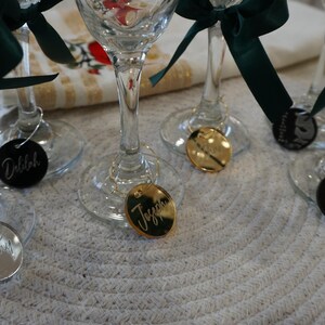 May include: Wine glasses with green ribbon bows and personalized name tags. Some tags are gold, some are black, and some are silver. The names are written in cursive script. The glasses are on a white, textured surface.