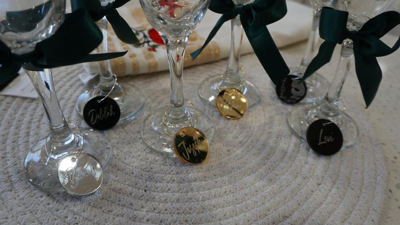 May include: Wine glasses with green ribbon bows and personalized name tags. Some tags are gold, some are black, and some are silver. The names are written in cursive script. The glasses are on a white, textured surface.