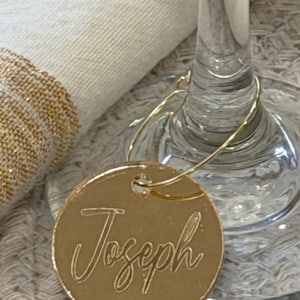 May include: A gold-colored, circular wine glass charm with the name "Joseph" in elegant script. The charm is attached to a wine glass, with a white cloth and gold-trimmed napkin in the background. The charm is designed to identify a guest's glass.