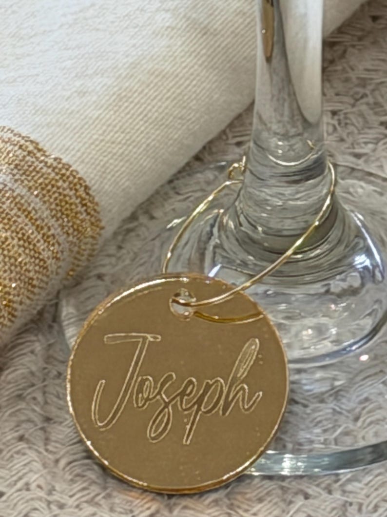 May include: A gold-colored, circular wine glass charm with the name "Joseph" in elegant script. The charm is attached to a wine glass, with a white cloth and gold-trimmed napkin in the background. The charm is designed to identify a guest's glass.