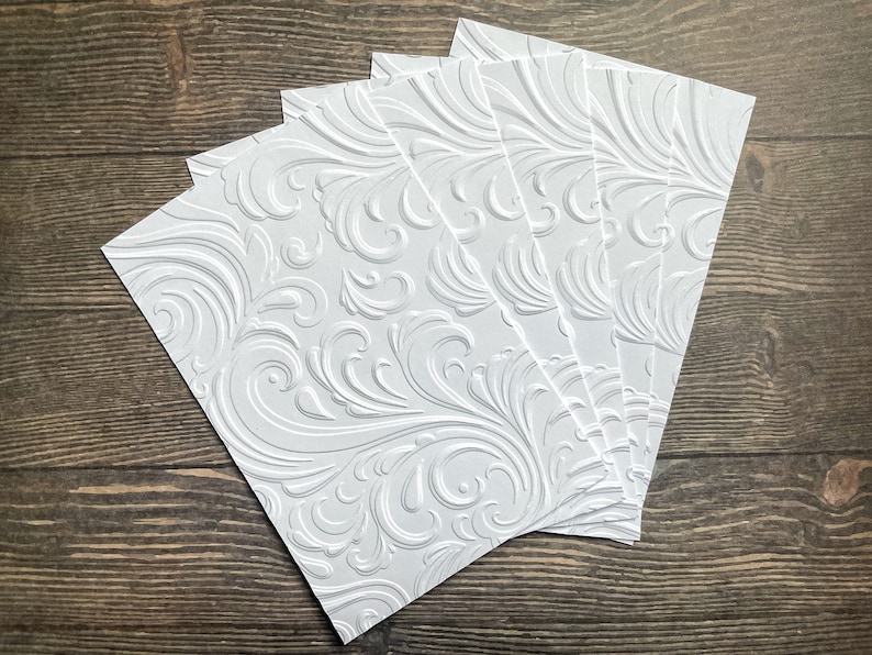 May include: Five white embossed cardstock sheets with a swirling, floral pattern. The sheets are stacked on top of each other.