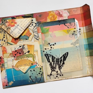 May include: A scrapbook featuring a collage of colourful paper, floral patterns, and butterfly images. The scrapbook has a layered design with pockets and flaps. The text "happy" is visible on a tag.