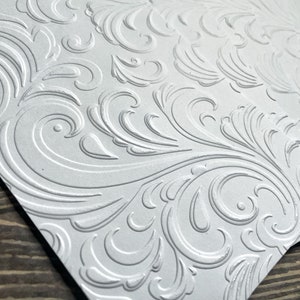 May include: A stack of white card stock with an embossed, intricate, swirling, floral pattern.