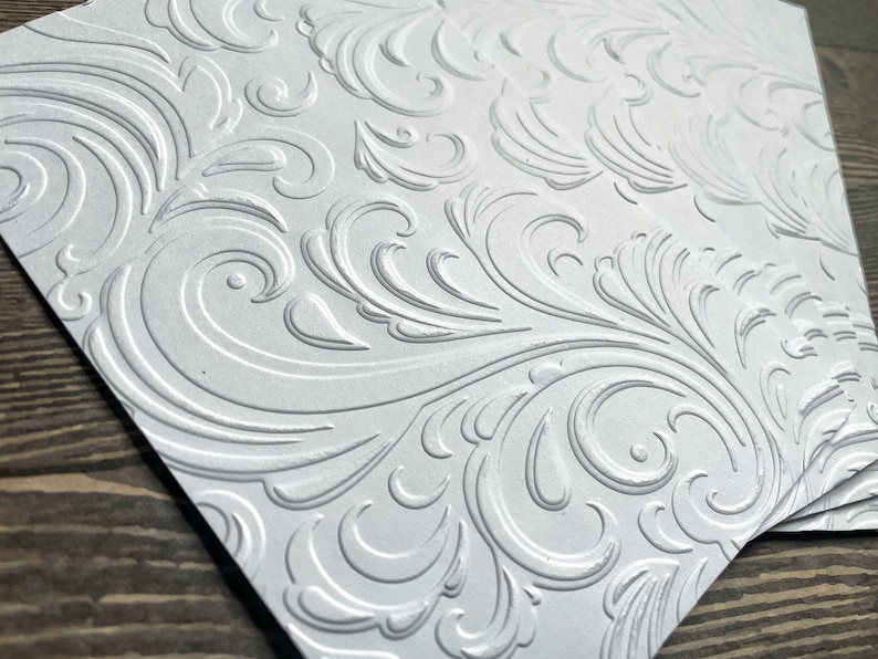 May include: A stack of white card stock with an embossed, intricate, swirling, floral pattern.