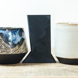 Könnte beinhalten: Drei Keramikobjekte auf einer Holzoberfläche. Eine Tasse mit Bergmotiv mit schwarzer, weißer und blauer Glasur, ein schwarzes, eckiges Objekt und eine weiße Tasse mit schwarzem Boden.