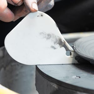 Peut inclure: Un outil blanc en forme de spatule est utilisé pour façonner un disque d'argile gris foncé sur un tour de potier. L'outil a un petit trou près de la poignée. La roue est noire et grise, avec un bord jaune visible.