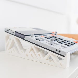 Può includere: Un campionatore di musica elettronica grigio chiaro con pulsanti e manopole neri e arancioni, poggiato su un supporto bianco stampato in 3D con un motivo geometrico a forma di montagna che inclina il dispositivo per un accesso più agevole.