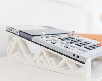 May include: A light grey electronic music sampler with black and orange buttons and knobs, resting on a white 3D-printed stand featuring a geometric mountain-like cutout pattern that elevates the device at an angled position for easier access.