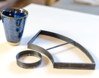 May include: A blue ceramic cup with a speckled finish and a light-coloured base, accompanied by a black circular ring-shaped cutter and a black fan-shaped cutter with a central divider, all arranged on a plain white surface.
