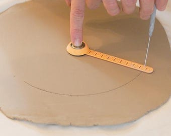 May include: A flat, light brown slab of clay being measured with an orange plastic pottery calliper tool. The tool features a central pivot point and a graduated scale with markings, used to scribe a circular outline into the surface of the clay.