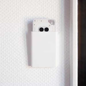 Könnte beinhalten: Ein weißes rechteckiges Gerät ist an einer strukturierten weißen Wand befestigt. Das Gerät hat ein rechteckiges weißes Gehäuse mit einer oben sichtbaren Kamerakomponente, die zwei schwarze kreisförmige Linsen aufweist. Die Wand hat ein subtiles, sich wiederholendes Muster.