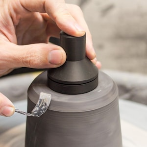 Könnte beinhalten: Hände eines Töpfers formen eine graue Keramikvase auf einer Töpferscheibe. Ein schwarzes Werkzeug wird verwendet, um die Form der Vase zu verfeinern. Die Vase ist auf der Scheibe zentriert, und die Hände des Töpfers führen den Prozess sorgfältig.