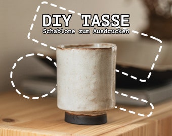 May include: A white ceramic mug with a dark brown rim and base. The mug is sitting on a wooden surface. The text "DIY TASSE Schablone zum Ausdrucken" is written in white on a white background with a dashed line border.