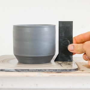 Puede incluir: Una maceta de cerámica gris con base negra se está midiendo con una herramienta de plástico negro. La maceta está sentada sobre una rueda giratoria gris y blanca.