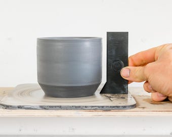 May include: A grey ceramic pot with a black base is being measured with a black plastic tool. The pot is sitting on a grey and white spinning wheel.